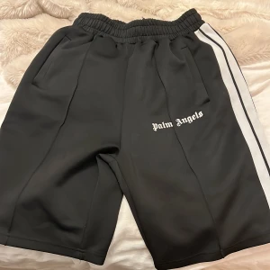 Svarta Palm Angels shorts med vita ränder - Svarta shorts från Palm Angels med vita sidoränder och logga broderad på ena benet. De har elastisk midja och är tillverkade i en mix av bomull och polyester. Sportig vibe och perfekt för dig som gillar streetwear.