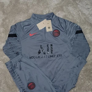 PSG långärmad träningsset från Nike - Paris Saint-Germain träningsset från Nike i mörkgrått med svarta och röda detaljer. Tröjan är långärmad med halv dragkedja och PSG-logga på bröstet. Byxorna har elastisk midja, PSG-märke och röd Nike Swoosh. Materialet är lätt och andas, perfekt för fotbollsträning.