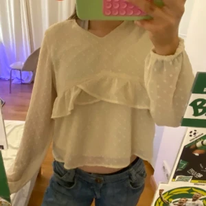 Kräm vit prickig blus med volang från Lindex - Supersöt vit blus från Lindex med diskret prickigt mönster och volangdetalj framtill. Blusen har v-ringning, långa ärmar med resår vid handlederna och en liten knapp i nacken. Tunt, lätt material som känns luftigt och fräscht.