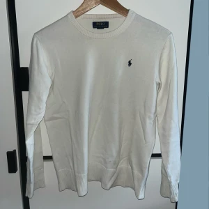Vit stickad tröja Polo Ralph Lauren - Klassisk vit tröja från Polo Ralph Lauren med rund halsringning och diskret broderad logga på bröstet. Tröjan har ribbade muddar vid ärmslut och nederkant, lång ärm och är tillverkad i mjuk bomull. Storlek XS passar ca 170cm