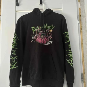 Rick and Morty svart hoodie - Svart hoodie med coolt Rick and Morty-tryck på bröstet och grönt tryck längs båda ärmarna. Tröjan har huva med snörning och en stor magficka. Perfekt för dig som gillar streetwear och vill sticka ut med en unik design.
