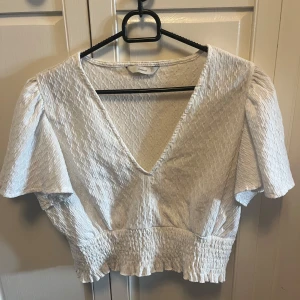 Vit blus med v-ringning från Only - Supersöt vit blus från Only med djup v-ringning, korta puffärmar och smockad midja. Blusen har en lätt struktur i tyget och är croppad med volangkant nertill. Perfekt för dig som gillar romantisk och trendig stil.