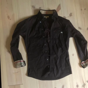 Svart  Burberry skjorta XS - Säljer en svart slim fit skjorta från Burberry London i storlek XS. Skjortan har klassiska rutiga detaljer på insidan av ärmsluten och kragen, knappar framtill och lång ärm. Tillverkad i bomull med en liten mängd elastan för extra komfort. Bara skriva vid frågor priset är inte hugget i sten.