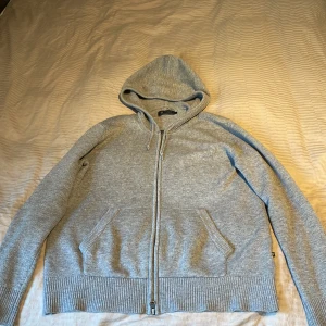 Grå stickad hoodie från Riley - Mysig grå stickad hoodie från Riley med dragkedja framtill, huva med snörning och ribbade muddar. Tröjan har fickor på magen och förstärkta armbågar. Perfekt för kyliga dagar och enkel att matcha med jeans eller joggers.