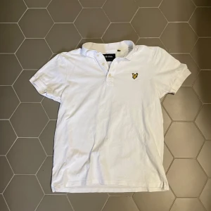Vit pikétröja från Lyle & Scott - Vit pikétröja från Lyle & Scott med klassisk krage och knappslå. Tröjan har korta ärmar och den ikoniska gula örnloggan broderad på bröstet. Tillverkad i bomull för en skön och luftig känsla.
