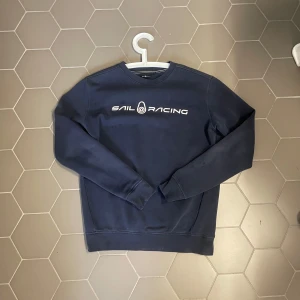 Marinblå Sail Racing sweatshirt - Snygg marinblå sweatshirt från Sail Racing med vit logga tryckt på bröstet. Tröjan har rund hals, långa ärmar och är gjord i mjuk bomullsmix. Perfekt för dig som gillar stilren och sportig look.