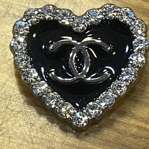  hjärtformad brosch med strass - Snyggt hjärtformat brosch i svart med silverfärgad logga. Runt kanten sitter glittrande strass-stenar som ger extra bling. Clip-on-modell som är lätt att bära och perfekt för dig som vill sticka ut.