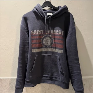 Saint Laurent  hoodie  - Snygg mörkblå hoodie från Saint Laurent med tryck i vitt och rött framtill. Tröjan har huva med snörning, känguruficka och långa ärmar. Materialet är mjukt och skönt, perfekt för chill dagar. Klassisk streetstil med coolt vintageinspirerat tryck.