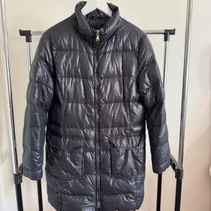 Svart pufferjacka Tommy Hilfiger M - Svart pufferjacka från Tommy Hilfiger i storlek M. Jackan har quiltad design, hög krage och två stora fickor framtill. Materialet är glansigt och syntetiskt, med blått foder på insidan. Perfekt för kalla dagar och har dragkedja hela vägen.