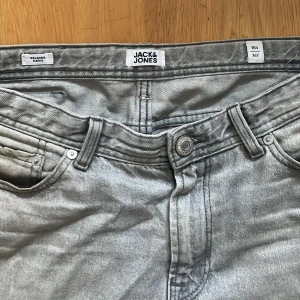 Grå jeans från Jack & Jones, straight fit - Snygga ljusgrå jeans från Jack & Jones i klassisk straight fit. Modellen har fem fickor, bälteshällor och knappgylf. Jeansen är tillverkade i bomull och har en cool tvättad look som passar perfekt till sneakers och hoodie.