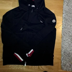 Svart Moncler hoodie med dragkedja - Svart hoodie från Moncler med vit och röd rand på huvan och muddar. Tröjan har snörning i huvan, Moncler-logga på ärmen och praktisk dragkedja framtill. Muddarna har snygga kontrastdetaljer i vitt och rött. Perfekt för dig som gillar stilrena streetwear-plagg.bnästsn oanvänd ..max 4 ggr har jag haft på mig den st M 