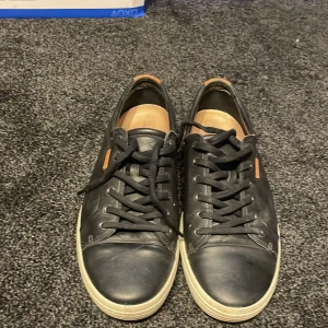 Svarta ECCO sneakers i skinn - Snygga svarta sneakers från ECCO i mjukt skinn med vita sulor och svarta skosnören. Klassisk rund tå och diskret ECCO-logga på sidan. Skorna har en stilren look och passar perfekt till jeans eller chinos.