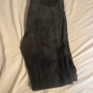 True Religion jeans med ormskinnsmönster - Säljer ett par svarta jeans från True Religion med unikt ormskinnsmönster. Modellen har raka ben och klassisk femficksdesign. Materialet är jeans och de har en snygg mörk ton som sticker ut. Perfekt för dig som vill ha något annorlunda i garderoben.