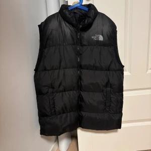 The North Face väst - Svart dunväst från The North Face i storlek XL. Västen har en hög krage, två sidofickor och en dragkedja framtill. Den är quiltad och har en stilren look med loggan tryckt på bröstet och ryggen. Perfekt för lager-på-lager under kyliga dagar.