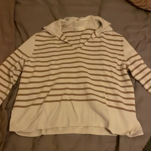 Beige och vit randig hoodie - Säljer en chill beige och vit randig hoodie. Tröjan har en mjuk huva, v-ringad hals och långa ärmar. Perfekt för dig som gillar avslappnad stil med lite preppy känsla. Materialet är skönt och lätt, passar bra till jeans eller mjukis.