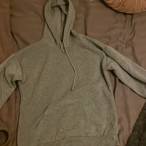 Grå hoodie med huva och snörning - En enkel grå hoodie med huva och snörning framtill. Tröjan har en klassisk känguruficka och långa ärmar. Perfekt för chill dagar och passar till det mesta. Materialet känns mjukt och bekvämt mot huden.