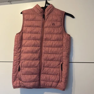 Rosa dunväst från Hangten - Snygg quiltad rosa dunväst från Hangten med hög krage och dragkedja framtill. Västen har två fickor med dragkedja och är perfekt för lager-på-lager. Den har en stilren, sportig look och är lätt att matcha med olika outfits.
