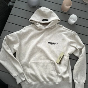 Grå Essentials Fear of God hoodie - Säljer en beige hoodie från Essentials Fear of God med svart logotyp på bröstet. Tröjan har huva, känguruficka och ribbade muddar. Perfekt för dig som gillar clean streetwear och vill ha en stilren look. Materialet är mjuk bomull och polyester.