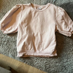 Ljusrosa sweatshirt från Gina Tricot - Supersöt ljusrosa sweatshirt från Gina Tricot med puffiga korta ärmar och ribbade muddar vid hals, ärmslut och nederkant. Mjuk insida och relaxed fit som ger en chill vibe. Perfekt att matcha med jeans eller kjol för en trendig look.