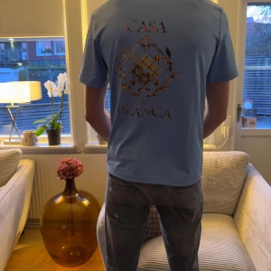 Blå Casa Blanca t-shirt med tryck - Snygg ljusblå t-shirt från Casa Blanca med stort grafiskt tryck i guld på ryggen och mindre tryck på bröstet. Klassisk rund hals och korta ärmar. Perfekt för dig som gillar stilrena plagg med coola detaljer.