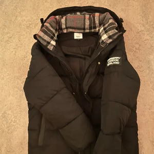 Svart pufferjacka från Burberry - Svart pufferjacka från Burberry med klassisk rutig insida i grått, vitt och rött. Jackan har huva, dragkedja och flera fickor med zip framtill. Stort Burberry London England-tryck på ärmen. Perfekt för kalla dagar och riktigt snygg streetstil.