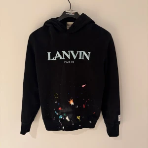 Svart Lanvin hoodie - Svart hoodie från Lanvin med stor logga framtill och coola färgstänk i olika nyanser på magen. Baksidan har trycket 'DÉPT. de la GALERIE' och en patch på ärmen. Mjuk bomullskvalitet och huva för extra chill vibe.