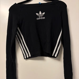 Svart croppad långärmad topp Adidas - Svart croppad långärmad topp från Adidas med klassiska vita ränder längs sidorna vid midjan och Adidas-logga framtill. Mjuk och skönt material💕