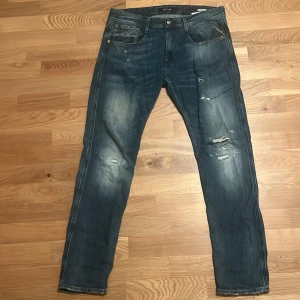 Replay Anbass blå jeans med slitningar - Säljer ett par Replay Anbass jeans i mörkblå tvätt med snygga slitningar och distressed detaljer på benen. Klassisk femficksmodell med knapp och dragkedja i gylfen. Jeansen har en slim passform och är tillverkade i stretchigt denim för extra komfort. Orgnal pris 1400, köpta för x-antal år sedan så dessa ser du inte på gatan 