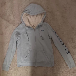 Ljusblå hoodie från Hollister med foder - Mysig ljusblå hoodie från Hollister med fluffigt foder i luvan och insidan. Tröjan har dragkedja, snörning i huvan, fickor fram och Hollister-logga på bröstet samt tryck längs ena ärmen. Perfekt för kyliga dagar.