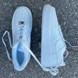 Nike Air Force 1 helvita sneakers - Klassiska Nike Air Force 1 sneakers i helvitt skinn med vit sula och snörning. Ikonisk design med perforerad tå, rund tå och Nike-logga på sidan. Perfekta för dig som vill ha en clean och tidlös look. Passar till allt och är superbekväma.