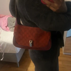 Röd axelväska från Gucci  - Snygg röd axelväska från Gucci med klassiskt GG-mönster och detaljer i skinn. Väskan har silverfärgat spänne framtill och en stilren, avlång form. Perfekt för dig som vill ha en ikonisk accessoar med lyxig känsla och tidlös design.
