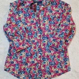 Blommig skjorta från GANT, strl 38 - Färgstark skjorta från GANT med blommigt mönster i rosa, blått, beige och inslag av grönt. Klassisk krage, knappar framtill och långärmad modell. Skjortan är gjord i mjuk bomull och har en normal passform som sitter snyggt på kroppen.