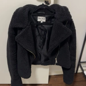 Svart teddyjacka från NA-KD - Mysig svart teddyjacka från NA-KD med oversized krage och grov dragkedja. Jackan är croppad och har en fluffig, fleece-liknande yta som ger en cool och avslappnad vibe. Perfekt för att hålla värmen under höst och vinter. Passar dig som gillar trendiga och bekväma plagg.