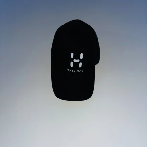 Haglöfs keps - Svart keps från Haglöfs med böjd skärm och vit logga framtill. Klassisk modell med justerbar rem baktill och ventilerande hål. Perfekt för dig som gillar stilren och sportig look. Materialet är troligtvis bomull eller polyester.