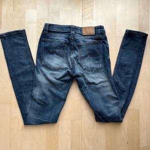 Nudie Jeans blå  W27 L34 - Snygga blå skinny jeans från Nudie Jeans med klassisk femficksdesign och coola slitningar. Jeansen har mörkblå tvätt med ljusare partier, brunt läderpatch med logga och broderade bakfickor. Perfekt passform och stretchigt jeanstyg.