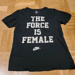 Svart Nike t-shirt med statementtryck - Svart t-shirt från Nike i bomull med vit text 'THE FORCE IS FEMALE' på framsidan och Nike-logga under. Klassisk rund hals och korta ärmar. Snygg och enkel passform, perfekt för dig som gillar att sticka ut med ett statement.