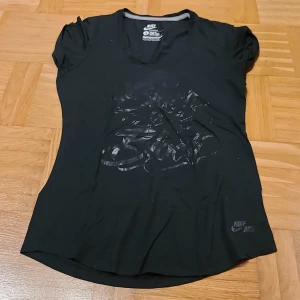 Svart Nike Air t-shirt loose fit - Svart t-shirt från Nike Air med loose fit och korta ärmar. Framsidan har ett coolt svart glansigt tryck i graffiti-stil och Nike Air-logga nere till höger. Materialet är mjuk bomull, perfekt för en chill och sportig look.