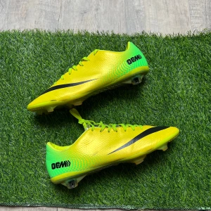 Nike fotbollsskor gul/grön med snörning - Säljer ett par grymma Nike fotbollsskor i neon-gult och grönt med svart logga och detaljer. Skorna har snörning, platt sula med dobbar och en sportig, aerodynamisk form. Ovandelen är i syntetmaterial och har en cool textur. Perfekt för dig som vill sticka ut på planen!