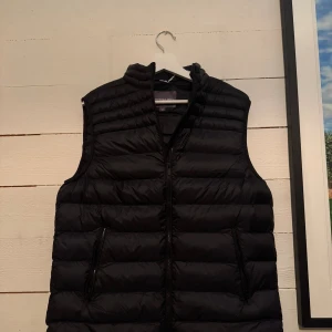 Svart dunväst från Lawrence Grey - Snygg svart dunväst från Lawrence Grey i storlek M. Västen har quiltad design, dragkedja framtill och två praktiska fickor med dragkedja. Perfekt att slänga över en hoodie för extra värme. Stilren look som funkar till många outfits, Helt ny aldrig använd.
