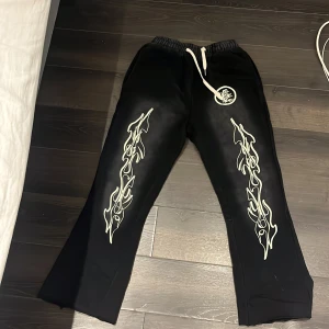 Svarta Hellstar mjukisbyxor med flames - Svarta mjukisbyxor från Hellstar med vita flames längs benen och logga på framsidan. Byxorna har snörning i midjan, en bakficka och är tillverkade i mjukt bomullsmaterial. Passformen är loose och benen är något utsvängda för en chill vibe.