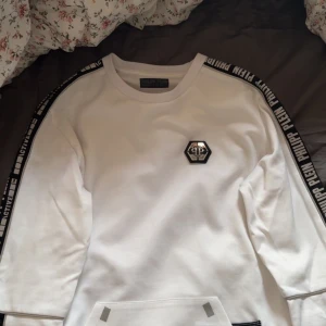 Vit sweatshirt från Philipp Plein - Säljer en clean vit sweatshirt från Philipp Plein med svarta logoband längs ärmarna och ett snyggt emblem på bröstet. Tröjan har lång ärm och rund hals, samt tryck på ryggen. Perfekt för dig som gillar streetwear med exklusiv vibe.