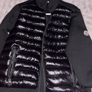 Svart dunjacka från Moncler - Svart dunväst från Moncler med glansig quiltad framsida och stickade svarta sidor. Västen har dragkedja framtill och två sidofickor med dragkedja. Moncler-logga på vänster ärm ger en exklusiv touch. Perfekt för lager-på-lager under kyliga dagar.