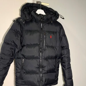 Ralph Lauren Svart pufferjaka  - Supersnygg svart pufferjacka från Polo Ralph Lauren med röd broderad logga på bröstet. Quiltad design, huva med snörning och flera dragkedjefickor. Varm, skön och har en clean, sportig vibe – perfekt för kalla dagar!