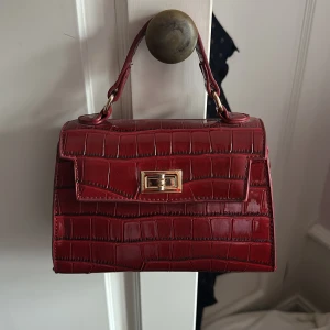 Röd handväska med krokodilpräglad yta - Snygg röd handväska med krokodilpräglad yta och guldfärgat lås. Väskan har ett stilrent, boxigt format och ett kort handtag med metallfästen. Insidan är svart och väskan stängs med ett vridlås i guld. Perfekt för dig som gillar statement-accessoarer. Skit snygg men kommer inte till användning längre pga har en annan väska ☺️‼️ 