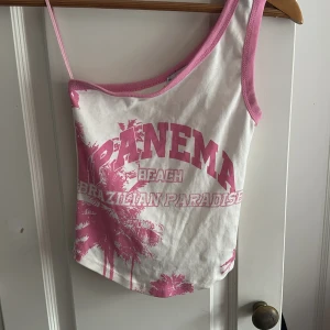 Vit och rosa oneshoulder topp Bershka - Säljer en vit och rosa oneshoulder topp från Bershka med tryck 'Ipanema Beach Brazilian Paradise' och palmmotiv. Toppen har rosa kantband och en smal axelrem på ena sidan. Perfekt för sommarvibbar och strandhäng. Skit snygg och gullig, kommer inte till användning ☺️ Använt endast vid köp ‼️☺️