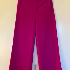 Fuchsia Sini vida byxor från Rodebjer S - Eleganta, fuchsiafärgade Sinibyxor med vida ben. Storlek S men jag  upplever dem större och med längre gren. Byxorna har en knappstängning med dekorativ knapp och praktiska fickor i sidorna. Obs uppfållade hos skräddare. Se mått på bilder 5-10! Ytterbenslängd 99,5 cm, innerbenslängd 65 cm, grenlängd fram från midja till grensöm 36,5 cm, grenlängd bak från midja till grensöm 43,5 cm. Midja 79 cm.