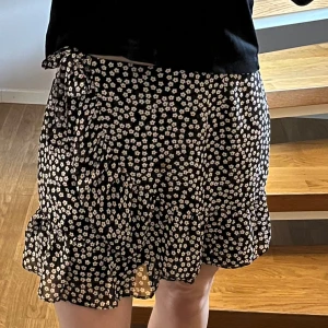 Blommig omlottkjol från Vintage Dressing - Supersöt svart kortkjol från Vintage Dressing med vita små blommor och röda detaljer. Kjolen har volangkant nertill och knytband i midjan för en omlott-look. Storleken är S/M men passar S bäst🩷