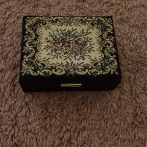 Broderad smyckesask med blommor - En unik smyckesask med broderat blommönster i beige, grönt och rosa på locket. Insidan är klädd i starkt orange tyg med fack för ringar och andra smycken. Perfekt för att hålla ordning på dina favoritsmycken.