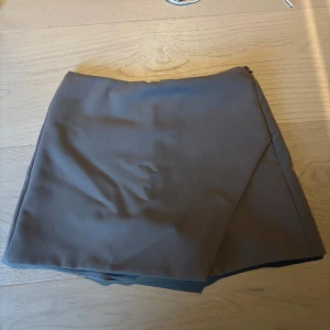 Grå omlottshorts från Zara XS - Stilrena grå/beiga shorts från Zara i omlottmodell med diskreta fickor bak. Klassisk design med hög midja och dragkedja i sidan. Perfekta för en clean och snygg look. Materialet känns mjukt och har en lätt stretch.
