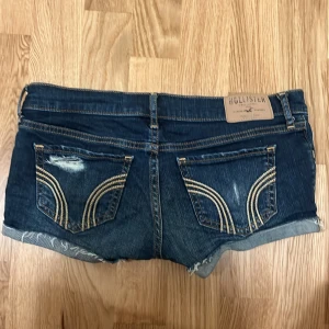 Mörkblå jeansshorts från Hollister - Snygga mörkblå jeansshorts från Hollister med slitna detaljer och uppvikta benslut. Shortsen har klassiska bakfickor med guldfärgad broderad söm och låg midja. Perfekta för en trendig y2k look. Dragkedjan är trasig men det går säkert att fixa och det är inget som syns när man har på sig dom.
